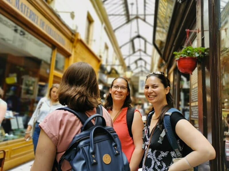 Trois femmes souriantes discutent dans un passage couvert à l’ambiance rétro, avec des vitrines de part et d’autre.
