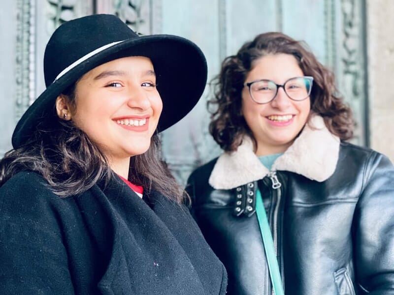 Deux amies souriantes posent devant une porte ancienne, l’une portant un chapeau noir, l’autre des lunettes.