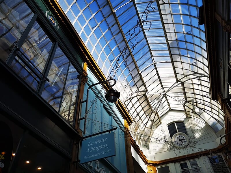 Verrière en verre d’un passage couvert parisien, laissant passer la lumière du jour, avec une horloge en fond.