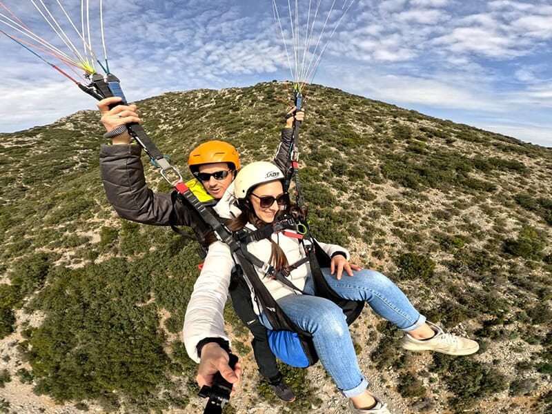 Deux personnes en tandem lors d’un vol en parapente au-dessus d’une colline boisée, souriantes et portant des casques.
