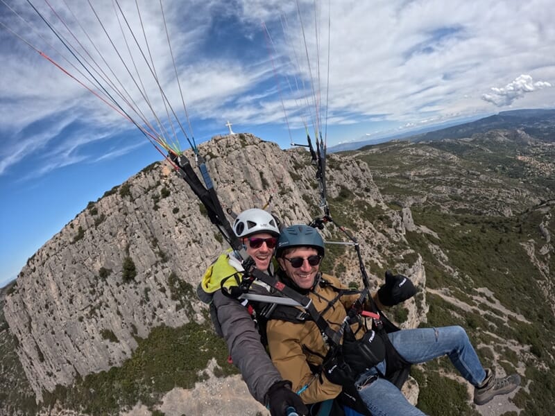 Deux hommes en parapente biplace volent au-dessus d’une falaise rocheuse avec une grande croix blanche au sommet, sous un ciel bleu partiellement nuageux.