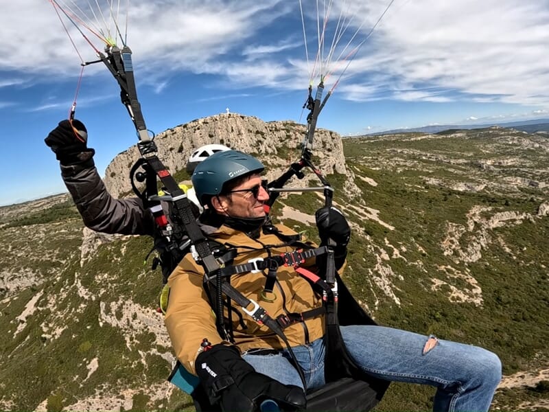Homme en veste marron et casque bleu assis en parapente, survolant un paysage montagneux verdoyant avec des formations rocheuses.