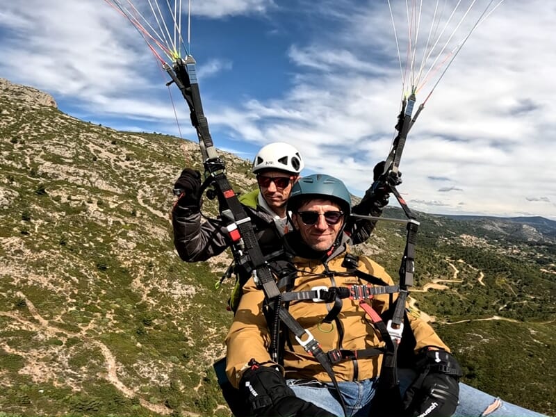 Deux hommes en vol en parapente, harnais visibles, ciel partiellement couvert et relief montagneux en arrière-plan avec chemins sinueux.