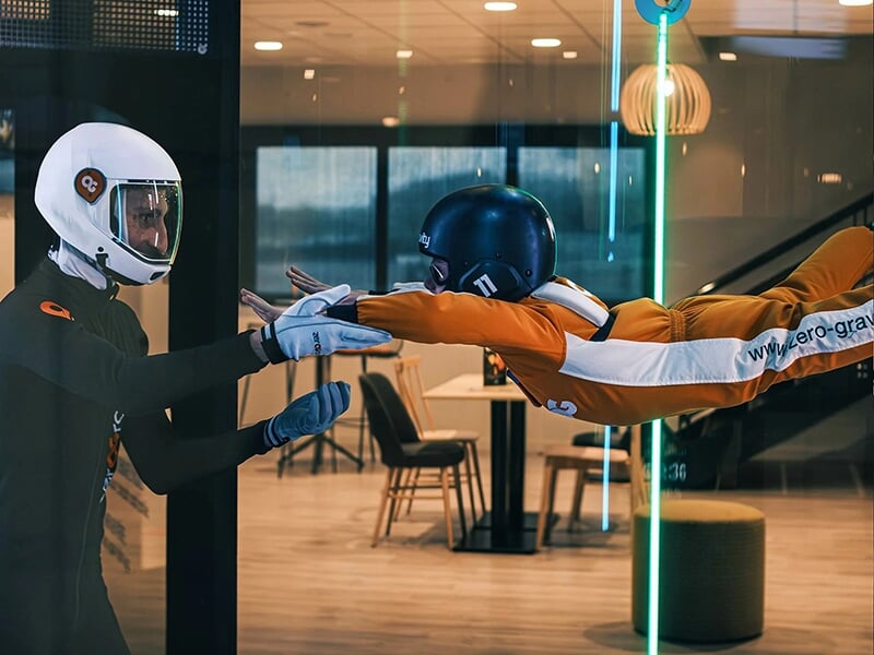 Personne en combinaison orange volant horizontalement dans un simulateur de chute libre, aidée par un instructeur en noir.