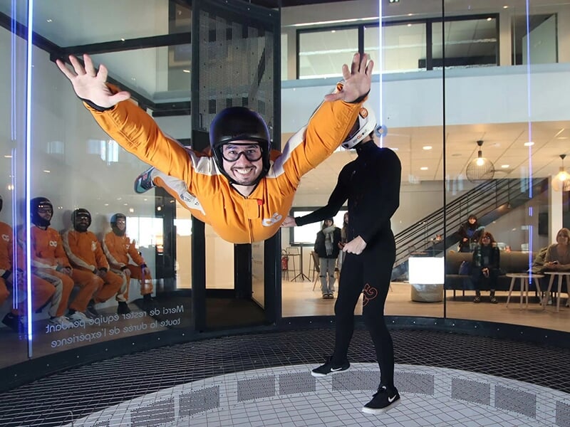 Homme souriant en plein vol dans un tunnel de vent, les bras écartés, accompagné d'un instructeur.