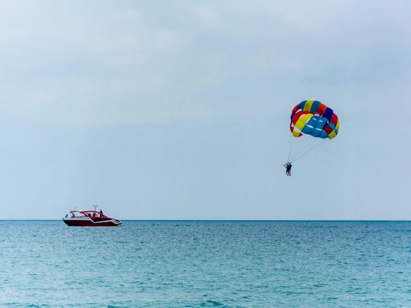 Un parachute multicolore tracté par un bateau rouge et blanc flotte au-dessus de la mer calme sous un ciel nuageux.