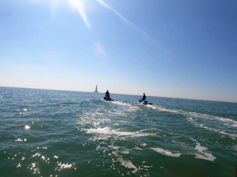 Deux personnes en jet-ski naviguent sur une mer calme sous un ciel bleu, laissant des sillages blancs derrière elles, avec un phare visible à l’horizon.