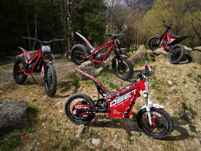 Quatre moto trial électriques disposées sur un sol rocheux avec un peu d'herbe et en fond la montagne et les arbres
