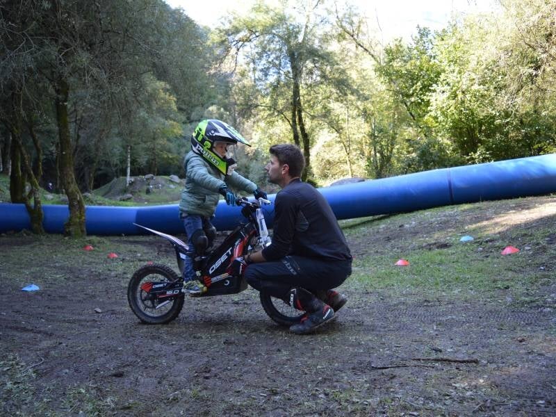 Dans une clairière, un enfant sur une moto trial électrique et son moniteur accroupi devant lui en train de lui parler