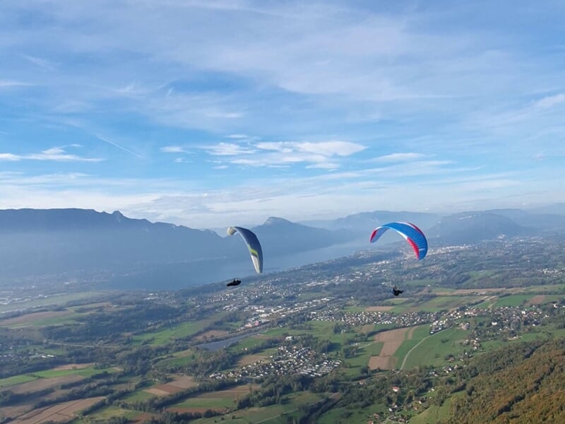 Deux parapentistes volent au-dessus d'un paysage vallonné avec un lac et des montagnes sous un ciel bleu clair.