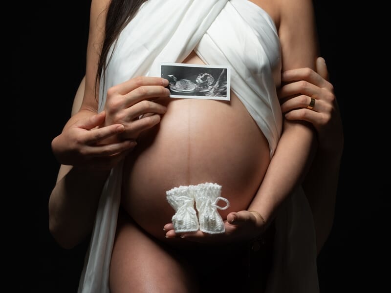 Photo d'une femme enceinte de face sur fond noir avec une photo de son échographie et des chaussons de bébé