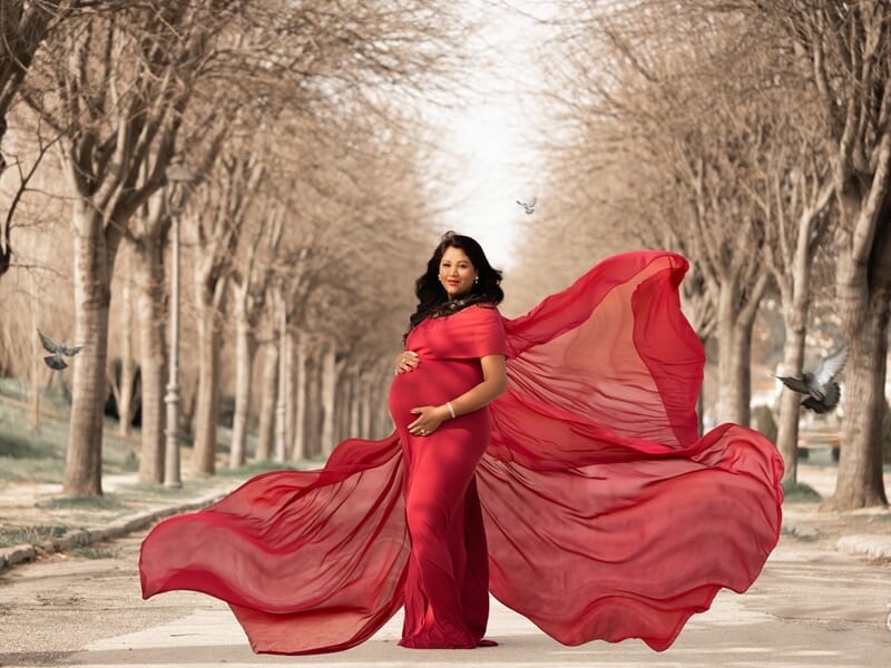 Photo d'une femme enceinte dans une allée d'arbres avec une robe rouge qui vole au vent