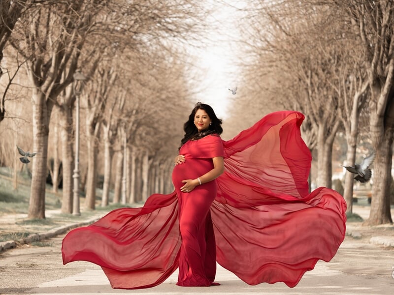Photo d'une femme enceinte dans une allée d'arbres avec une robe rouge qui vole au vent