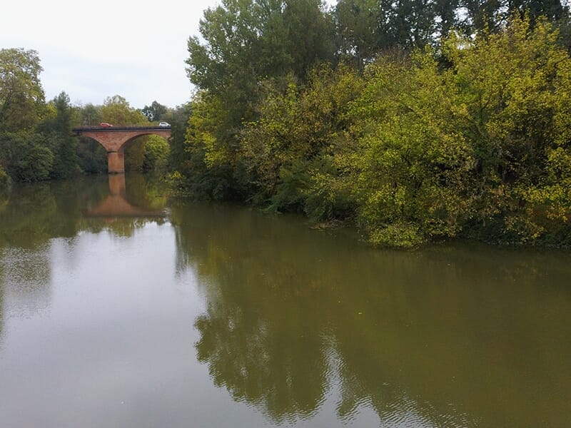 Pont en briques traversant une rivière entourée d’arbres, avec des voitures en circulation.