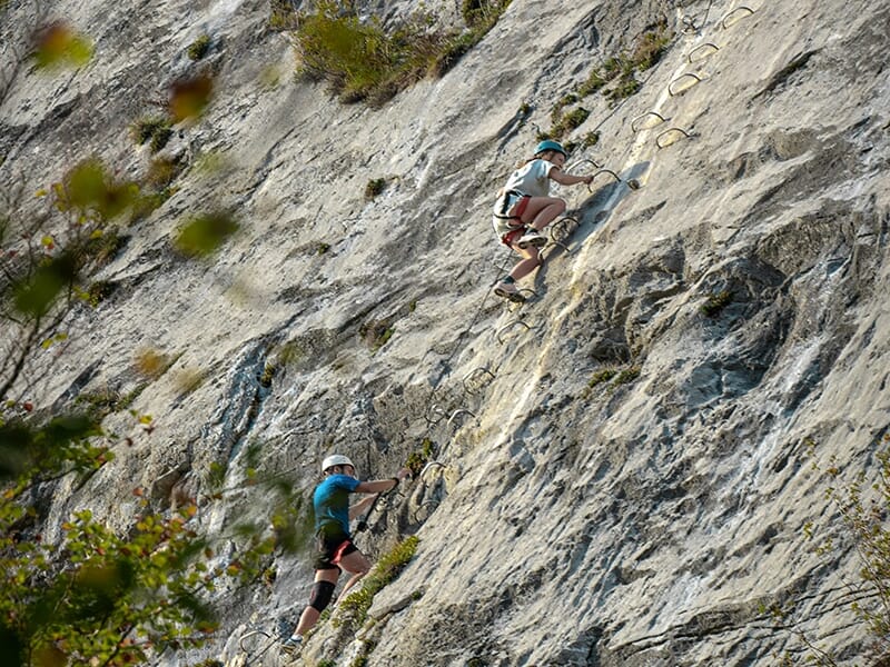 Deux personnes escaladant une via ferrata avec des barreaux métalliques sur une paroi inclinée.