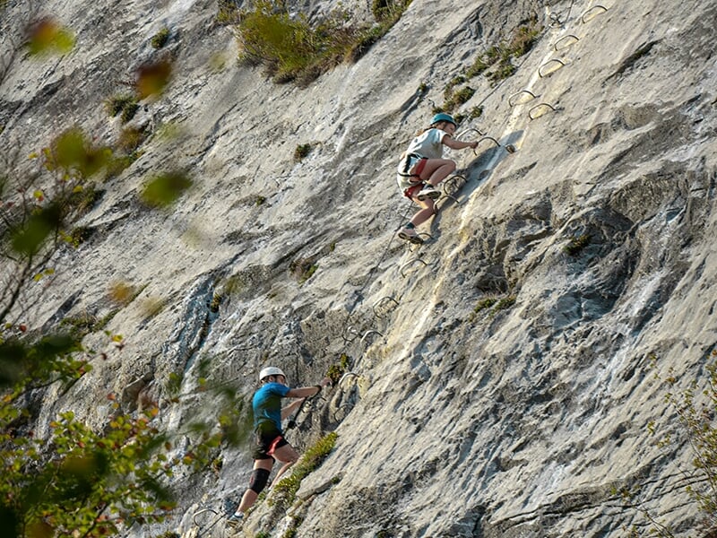 Deux personnes escaladant une via ferrata avec des barreaux métalliques sur une paroi inclinée.