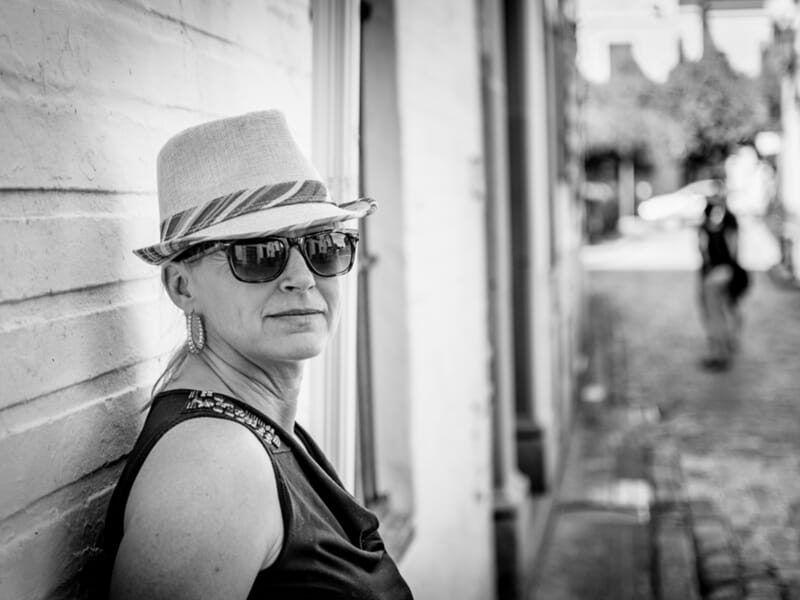 Portrait en noir et blanc d’une femme portant des lunettes de soleil et un chapeau, appuyée contre un mur en ville.