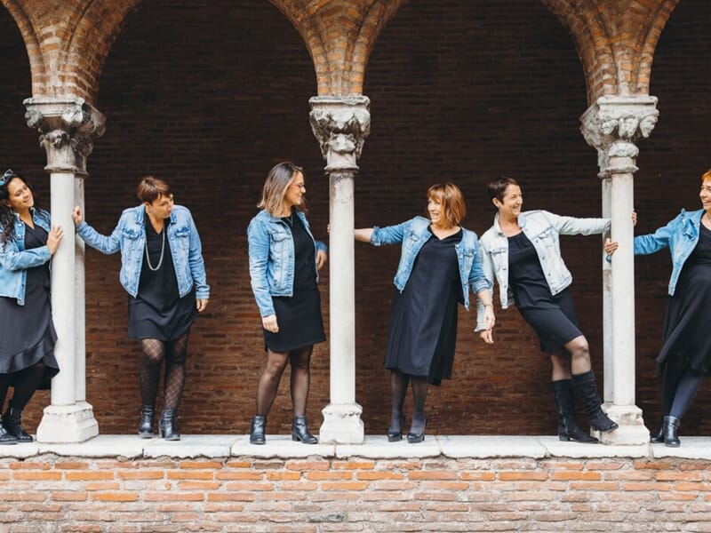 Six femmes en robe noire et veste en jean posent avec complicité entre les colonnes d’un cloître en briques.