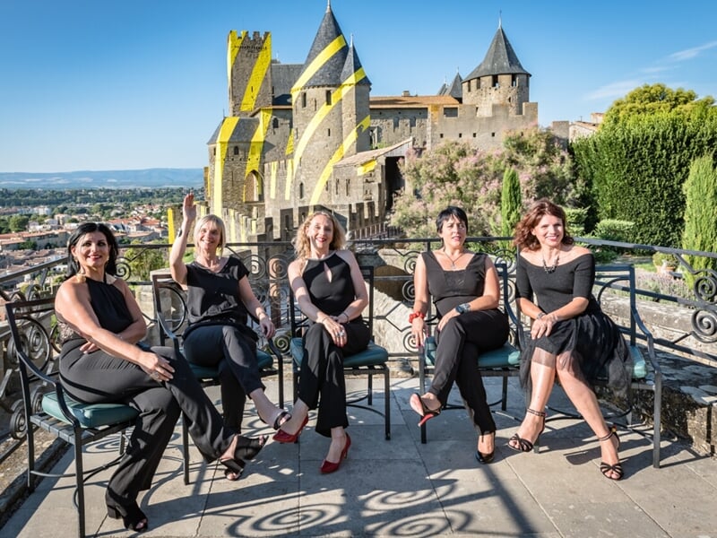 Cinq femmes élégamment vêtues posent sur une terrasse avec vue sur la cité médiévale de Carcassonne.