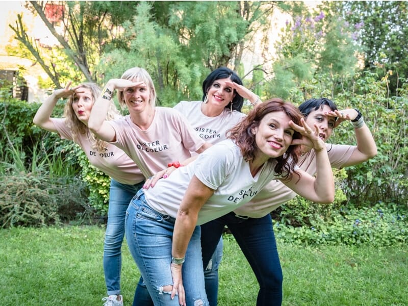 Cinq femmes en t-shirts assortis et jeans posent de façon amusante dans un jardin, mimant une posture de guet.