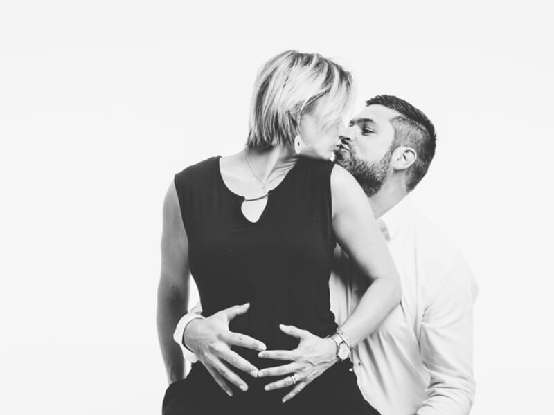 Portrait en noir et blanc d’un couple s’embrassant, l’homme pose les mains sur le ventre de sa compagne enceinte.