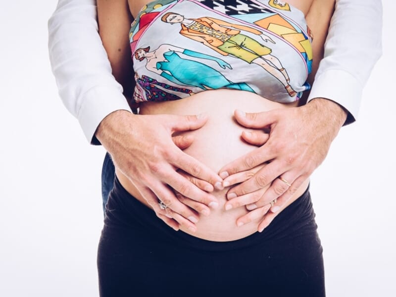 Mains d’un couple posées en forme de cœur sur le ventre rond de la future maman, vêtue d’un haut coloré.