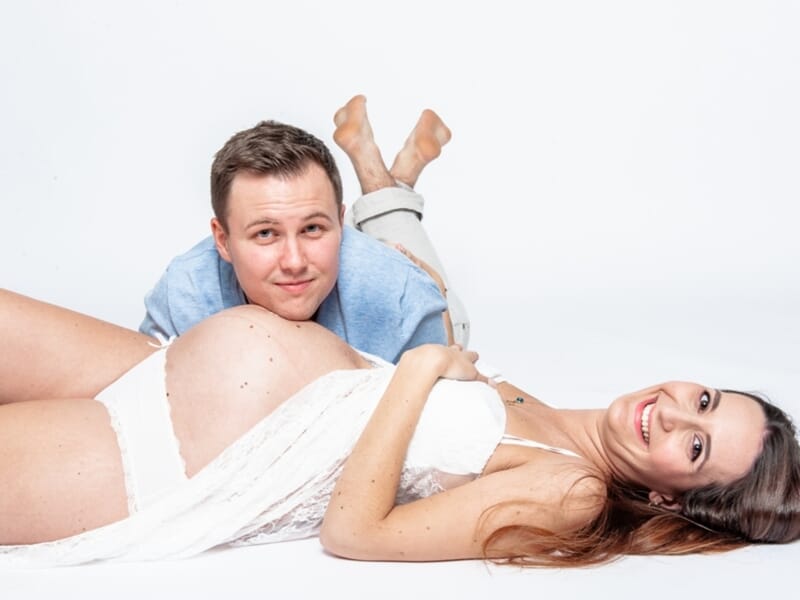 Femme enceinte allongée en lingerie blanche, souriante, son compagnon allongé derrière elle.