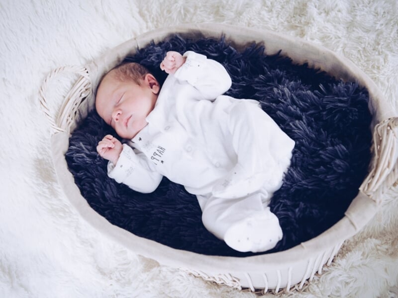 Bébé endormi en pyjama blanc dans un panier tapissé de fourrure noire, posé sur une couverture douce.