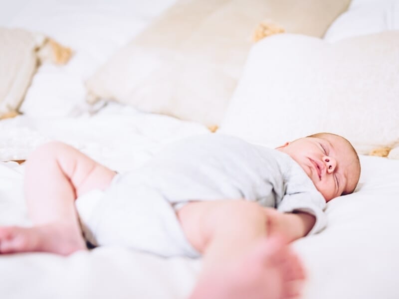 Nourrisson endormi sur un lit blanc entouré de coussins, en body gris clair.