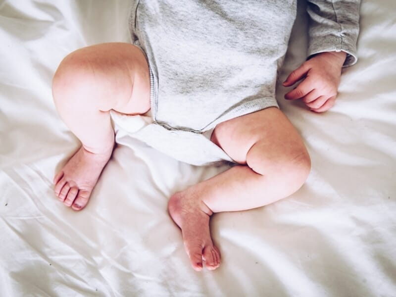 Gros plan sur les jambes potelées et les petites mains d’un bébé en body gris sur un drap blanc.