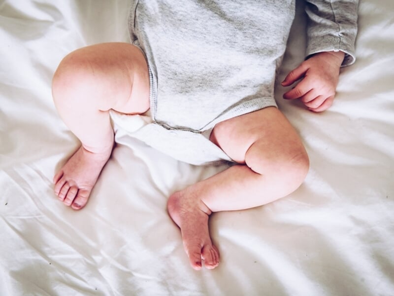 Gros plan sur les jambes potelées et les petites mains d’un bébé en body gris sur un drap blanc.