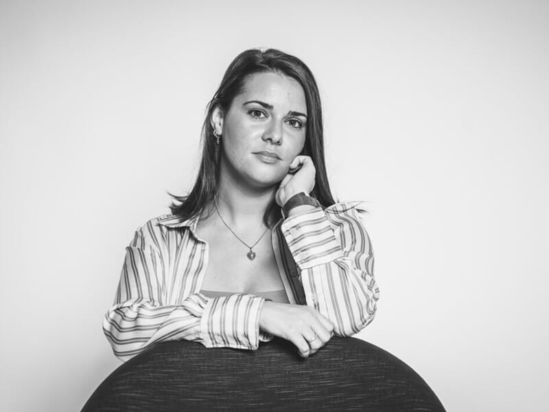 Portrait en noir et blanc d’une femme posant assise derrière un fauteuil rond, chemise rayée ouverte.