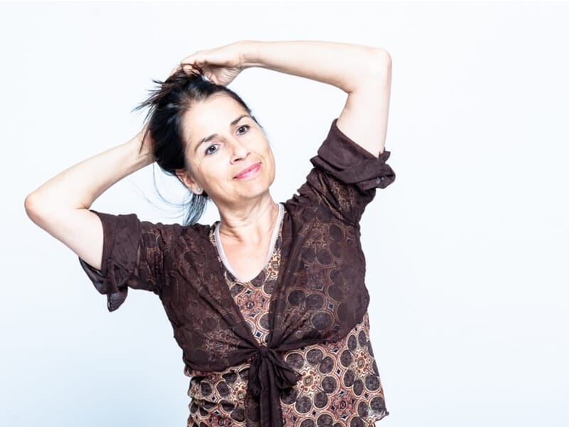 Portrait d’une femme souriante les bras levés, les mains dans les cheveux, posant sur fond clair.