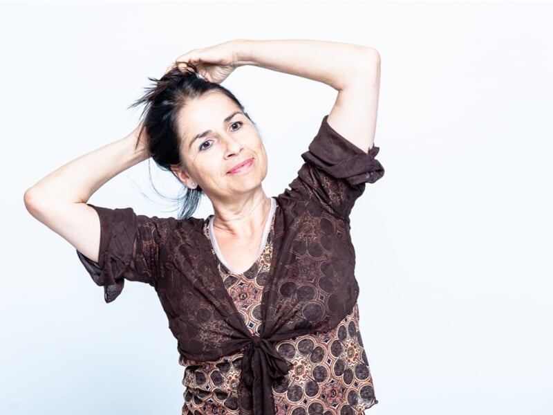 Portrait d’une femme souriante les bras levés, les mains dans les cheveux, posant sur fond clair.