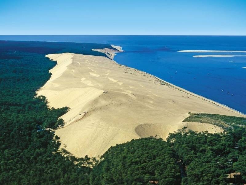 Vue aérienne sur la dune du Pilat, faite de sable jaune, entourée d'arbres verts et avec l'océan
