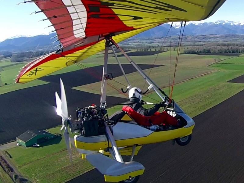 ULM pendulaire blanc et jaune avec une voile jaune et rouge, volant au dessus de champs verts et marron