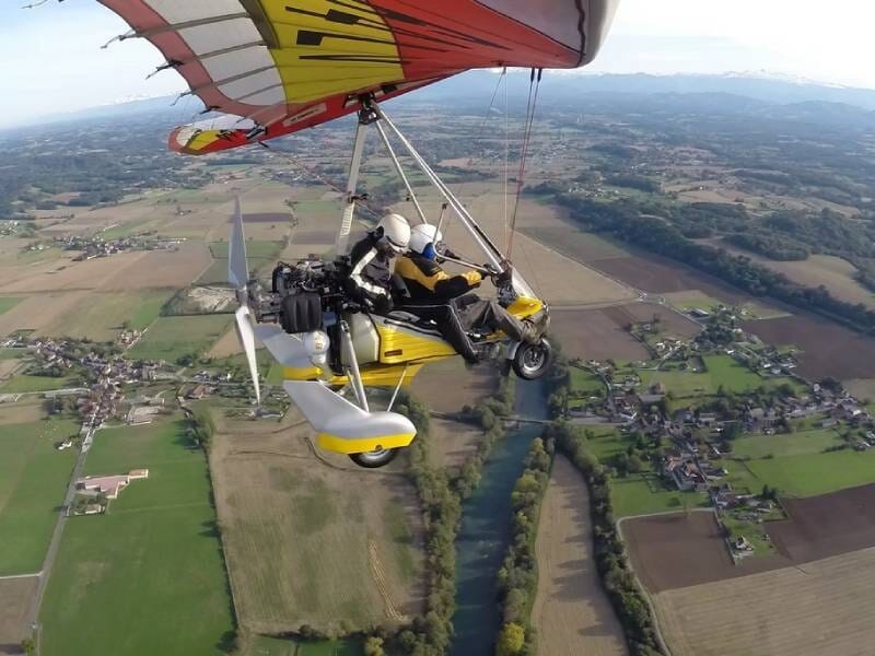 Un ULM pendulaire jaune et rouge en vol au-dessus des champs verts et marron