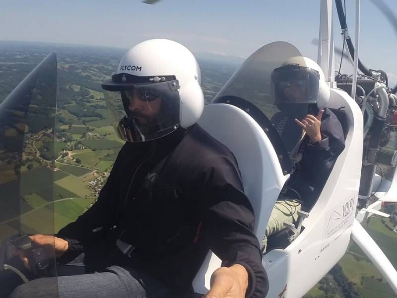 Un ULM autogire blanc en vol avec deux passagers qui portent des casques