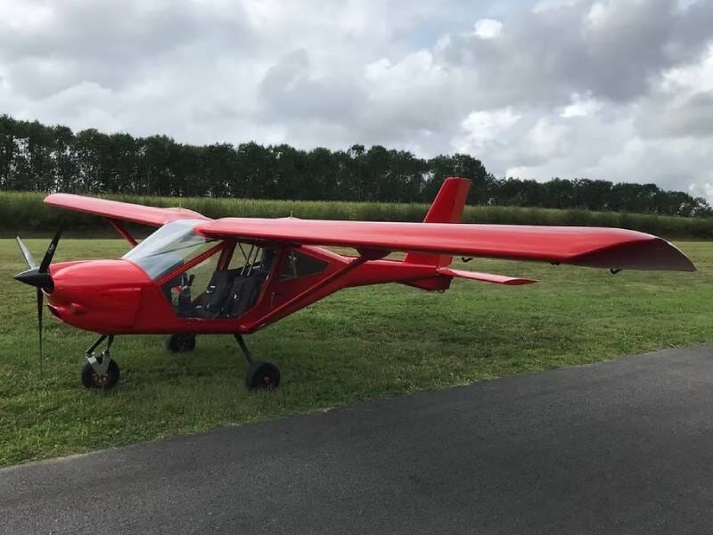 Avion ULM multiaxes rouge posé au sol, entre herbe et piste de décollage