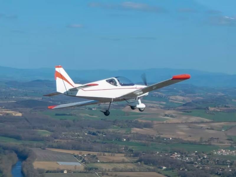 ULM multiaxes blanc et rouge en vol au-dessus des champs et d'un cours d'eau.