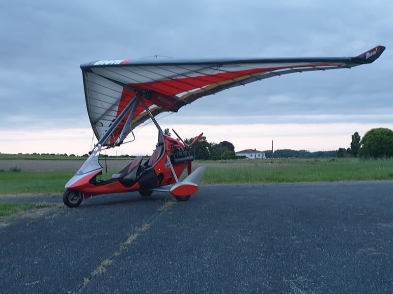 ULM pendulaire blanc et rouge posé sur une piste avec un champ d'herbe en fond.