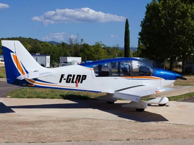 Avion ULM DR400 blanc et bleu posé au sol. Des arbres et le ciel bleu sont visibles en fond.