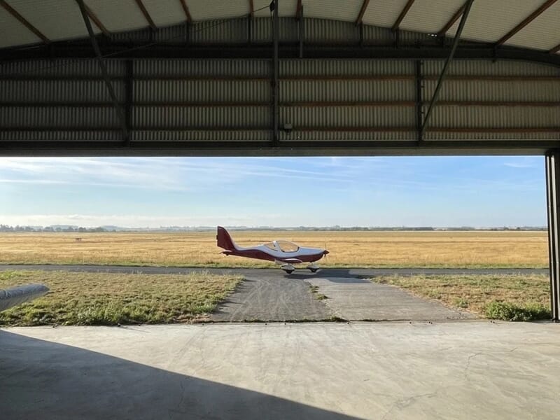 Avion rouge et blanc stationné devant un hangar ouvert avec une piste lors d'un vol en avion ULM à Lens.