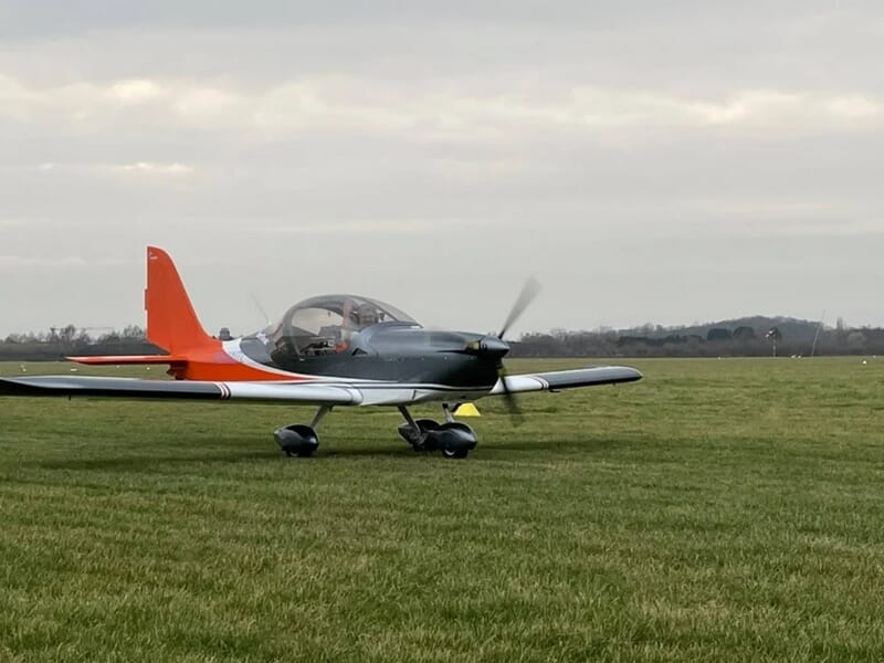 Petit avion gris et orange posé sur une piste d'herbe, avec un paysage rural lors d'un vol en avion ULM près de Lille.