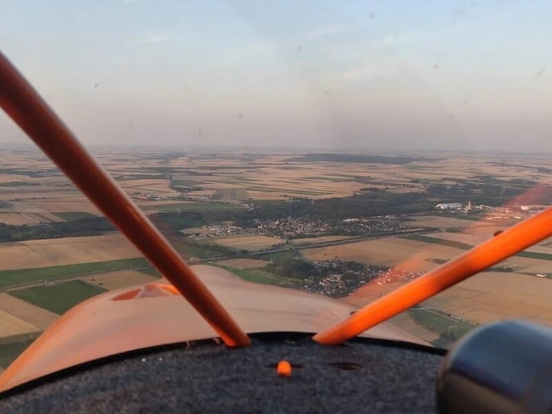 Vue aérienne depuis un ULM orange survolant des champs et un village à l'horizon lors d'un vol en autogire près de Reims.