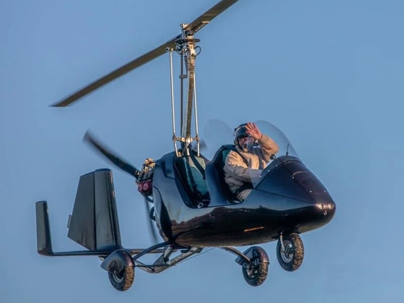 Gyrocoptère noir en vol avec un pilote saluant, lors d'un vol en autogire près d'Oloron-Sainte-Marie.