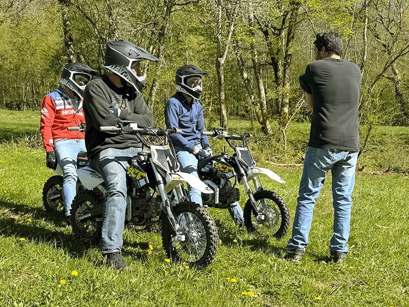 Trois jeunes en casque discutent avec un instructeur en plein air, assis sur des mini motos tout-terrain dans une clairière boisée.