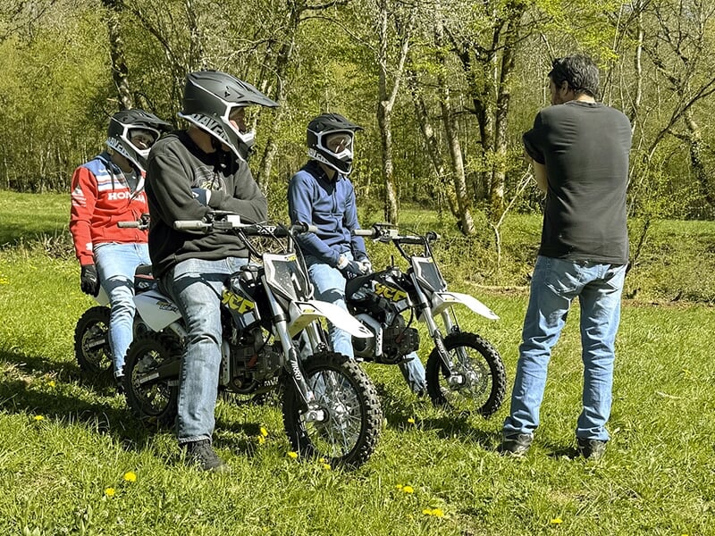 Trois jeunes en casque discutent avec un instructeur en plein air, assis sur des mini motos tout-terrain dans une clairière boisée.