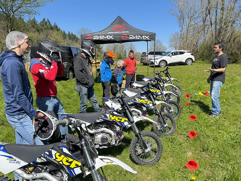 Participants debout à côté de leurs motos sous une tente marquée 24MX, écoutant un instructeur à l’extérieur.
