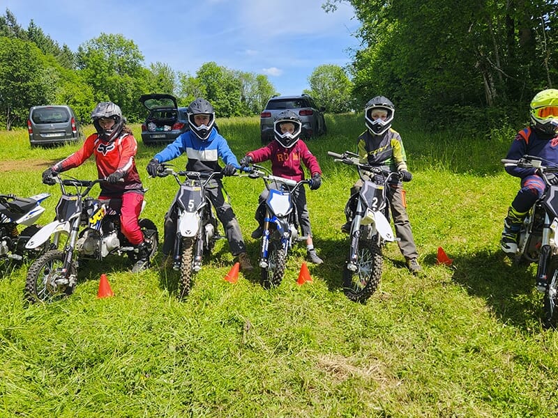 Cinq enfants alignés sur leurs motos dans une prairie, prêts pour une activité de groupe, entourés de voitures et d’arbres.